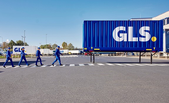 Équipe GLS sur site logistique traversant une zone sécurisée devant une semi-remorque bleue, centre de distribution et transport de colis.