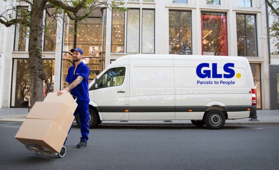 A GLS driver pushes packages on a dolly in front of a GLS van on a city street.